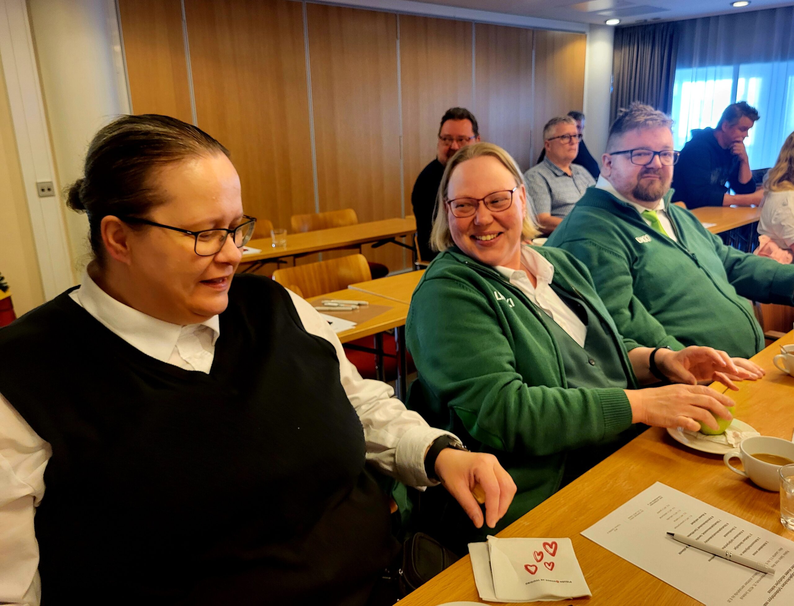 Laura Ahmio musta liivi päällä valkoisessa paidassa, Jonna Ahlsten vihreässä puserossa ja Riku Antikainen vihreässä puserossa ovat pöydän ääressä ja nauravat iloisina.