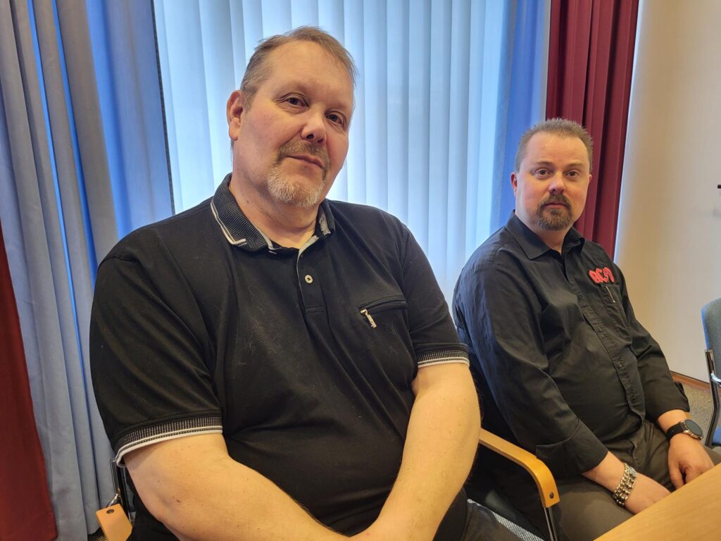 Kaksi miestä Jari Nevalainen ja Janne Ryynänen istuvat vierekkäin pöydän ääressä tyynen näköisinä. Taustalla sinertävät verhot.