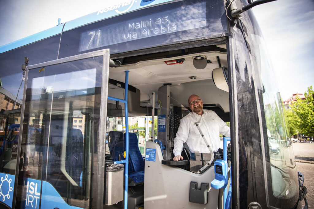 Kuvassa iloinen Pertti Piirinen bussin ohjaamossa. Kuva otettu bussin ulkopuolelta ja bussin ovet ovat auki.