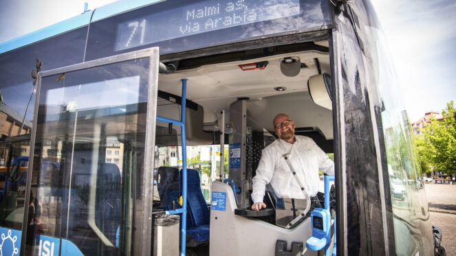 Kuvassa iloinen Pertti Piirinen bussin ohjaamossa. Kuva otettu bussin ulkopuolelta ja bussin ovet ovat auki.