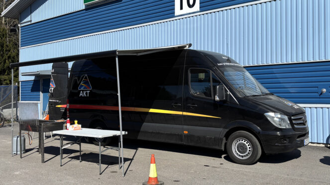 Kuvassa AKT:n tumma pakettiauto. Taustalla siniraidallinen rakennus