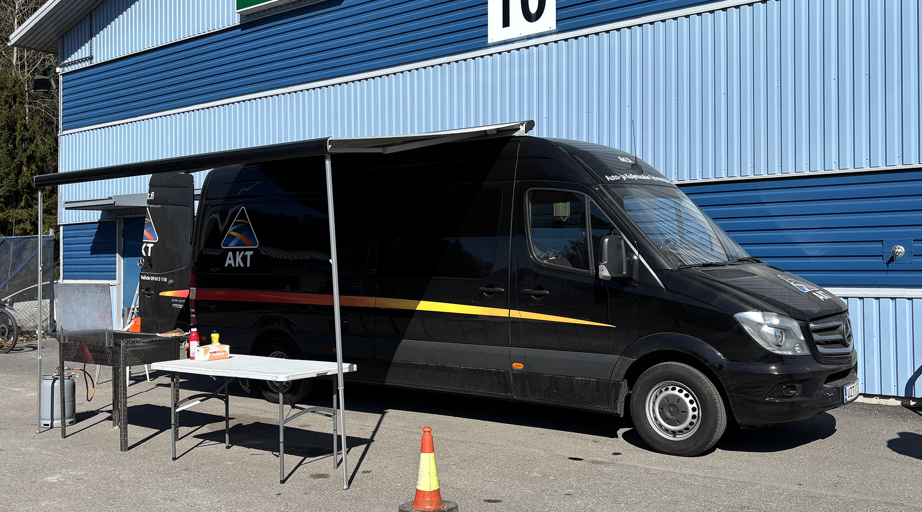 Kuvassa AKT:n tumma pakettiauto. Taustalla siniraidallinen rakennus