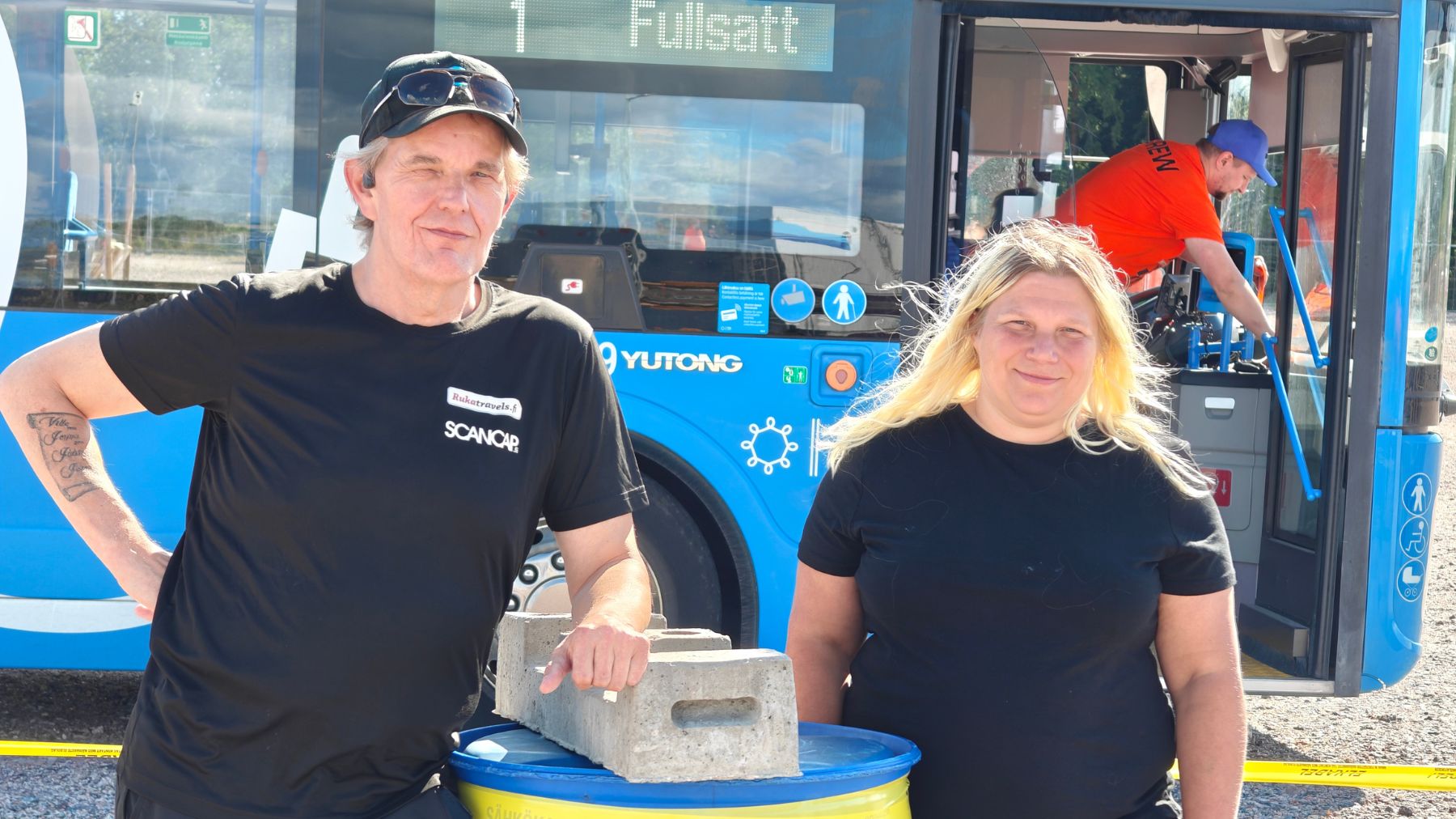 Mies ja nainen kuvassa nojaamassa tynnyriin. Molemmilla mustat t-paidat, miehellä lippis naisella vaaleat hiukset. Takana linja-auto.