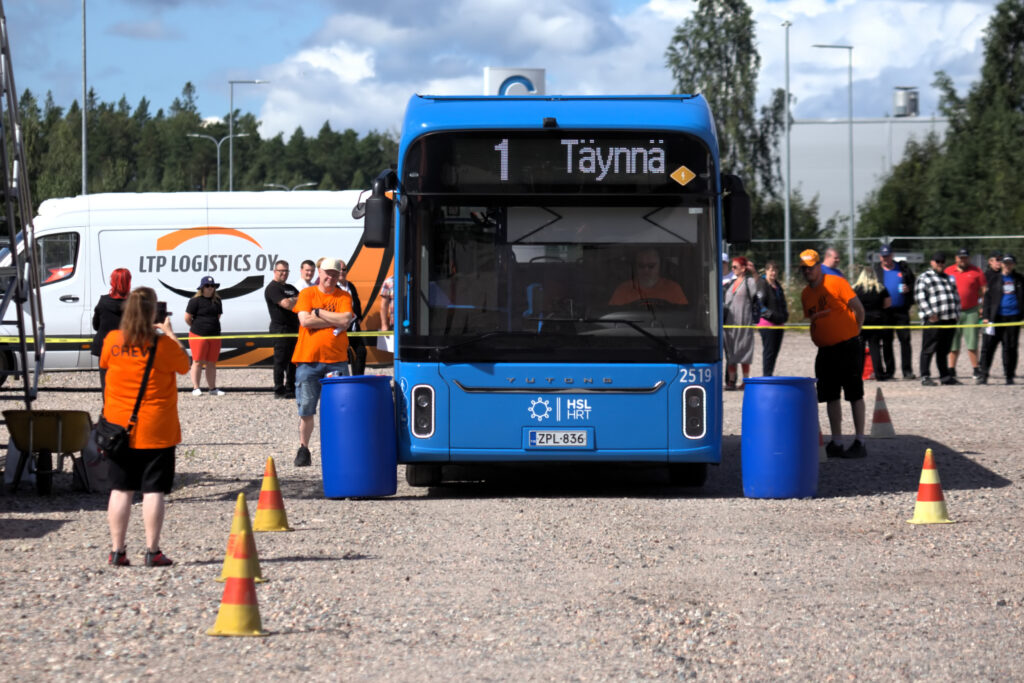 Linja-auto kahden sinisen tynnyrin välissä. Ympärillä toimitsijoita seuraamassa, miten tarkkuusajo sujuu.