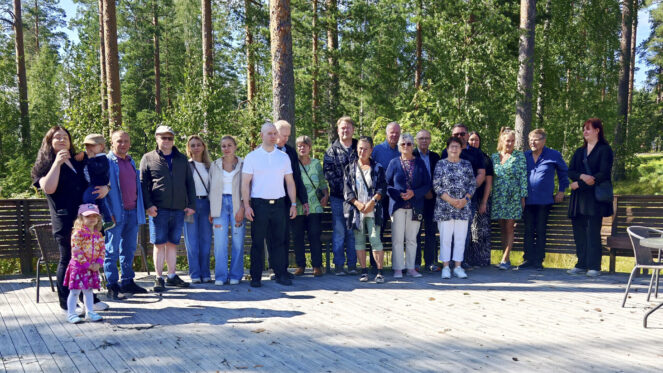 Kuvassa joukko eri ikäisiä ihmisiä poseeraa kesäisissä maisemissa.
