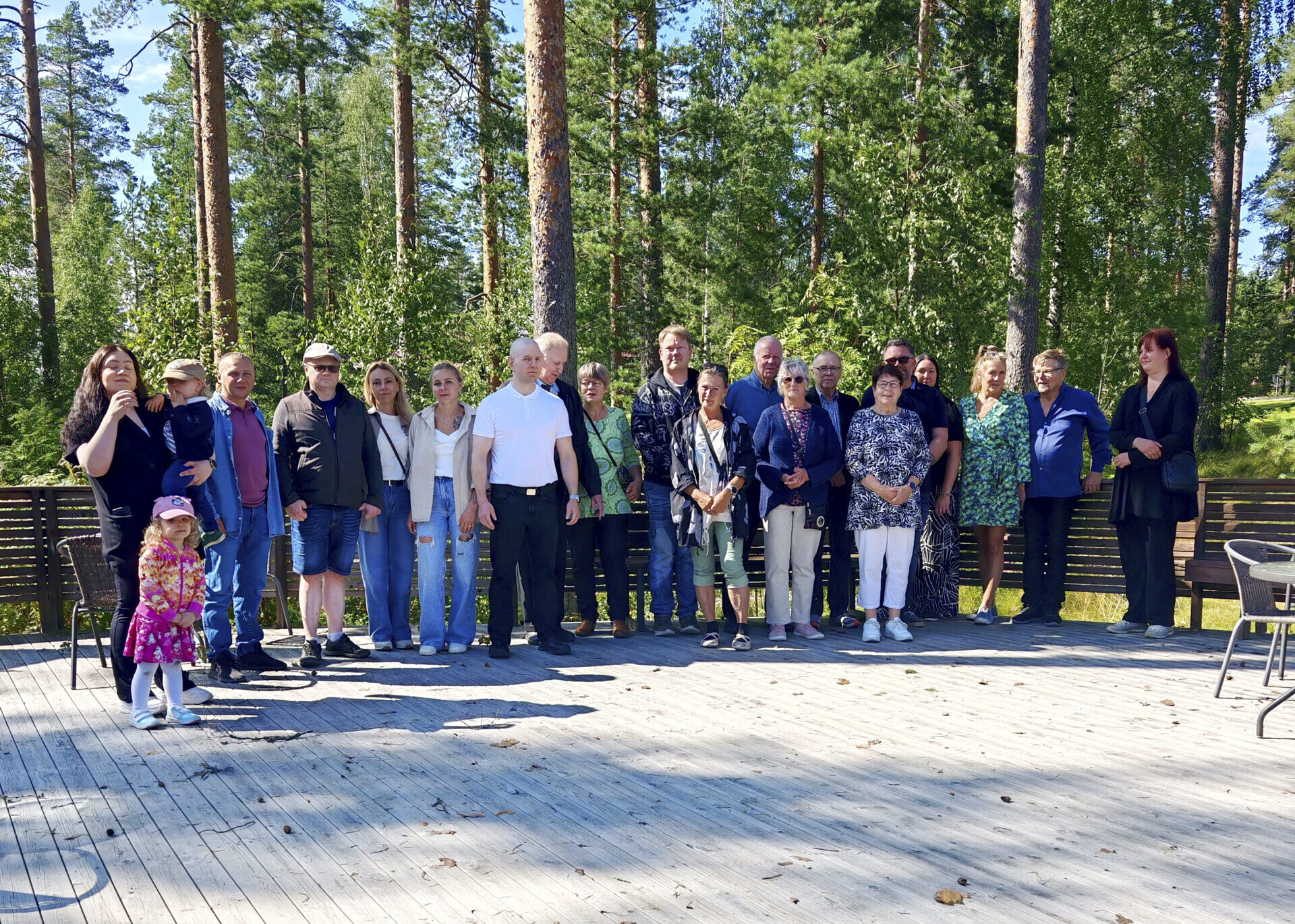 Kuvassa joukko eri ikäisiä ihmisiä poseeraa kesäisissä maisemissa.