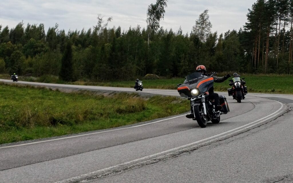 Kuvassa moottoripyöräilijöitä ajamassa tienmutkassa. Taustalla puita ja pilvinen sää.