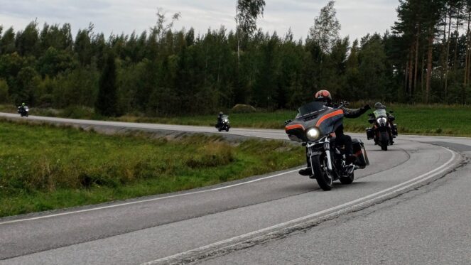Kuvassa moottoripyöräilijöitä ajamassa tienmutkassa. Taustalla puita ja pilvinen sää.
