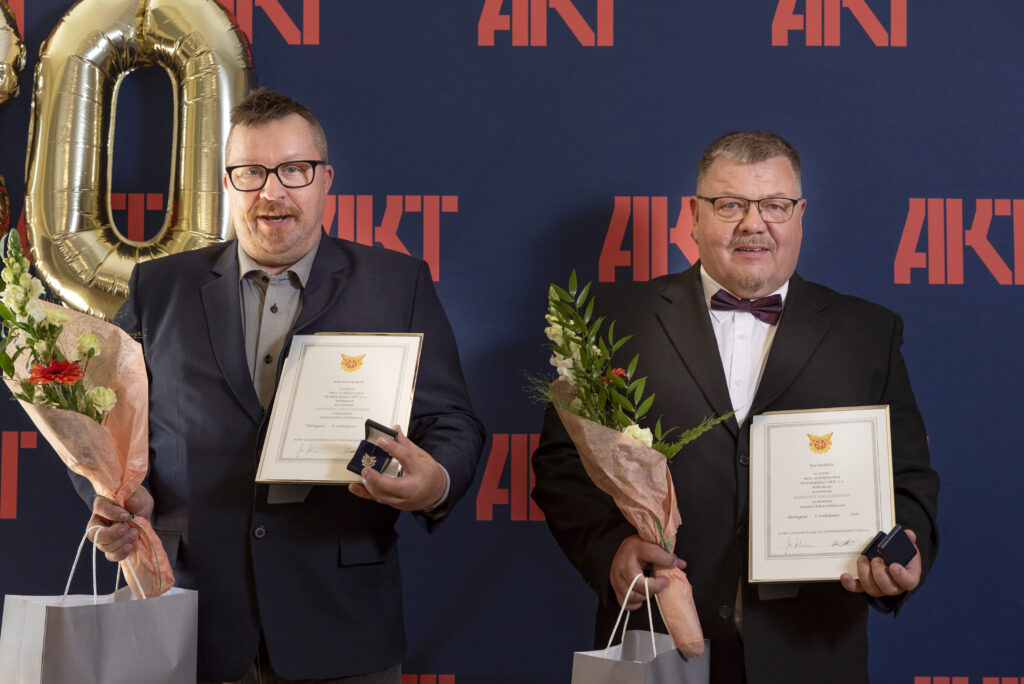 Kuvassa Jani Korpela ja Tero Jaakkola poseeraavat kukkien ja ansiomerkkien kanssa AKT-plakaatin edessä juhlallisissa vaatteissa.