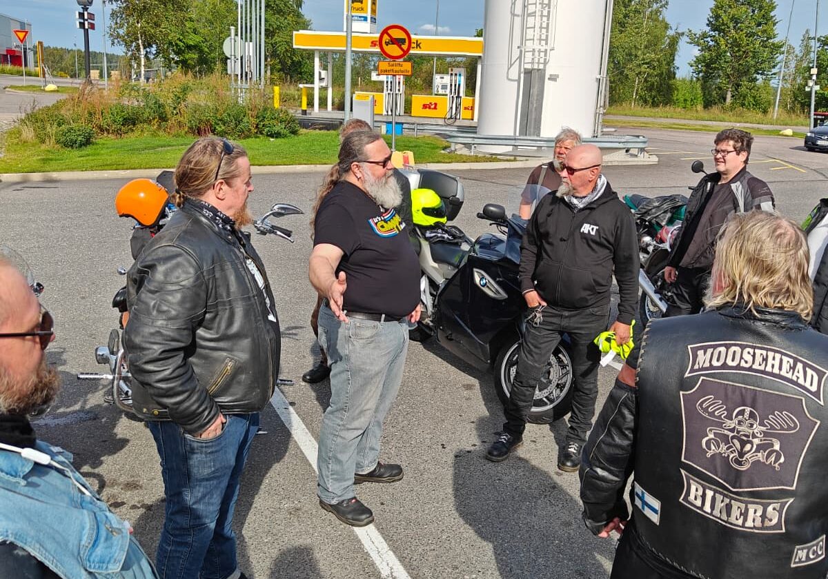 Kuvassa moottoripyöräilijähenkiset ihmiset seisovat parkkipaikalla katseet toisiinsa kääntyneinä.