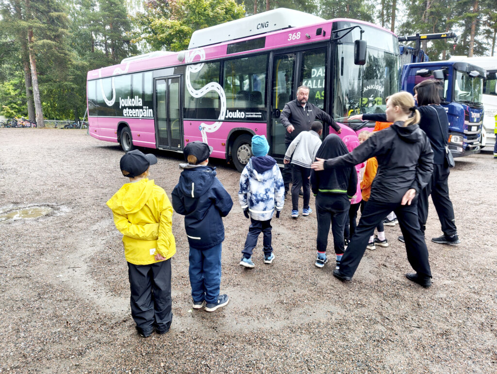 Lauma lapsia jonottaa kahden aikuisen avustamana vaaleanpunaiseen linja-autoon.