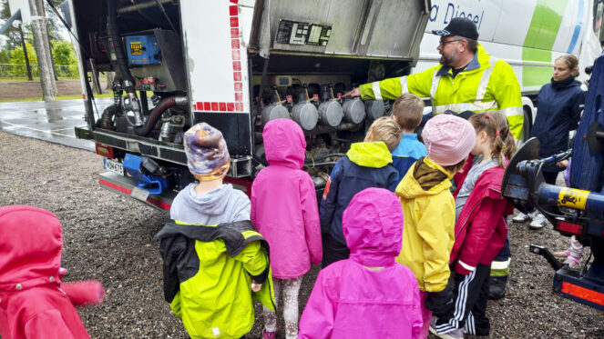 Lauma värikkäisiin vaatteisiin pukeutuneita lapsia seisoo säiliöauton edessä, jota esittelee henkilö neonvihreässä työtakissaan ja lippiksessään.
