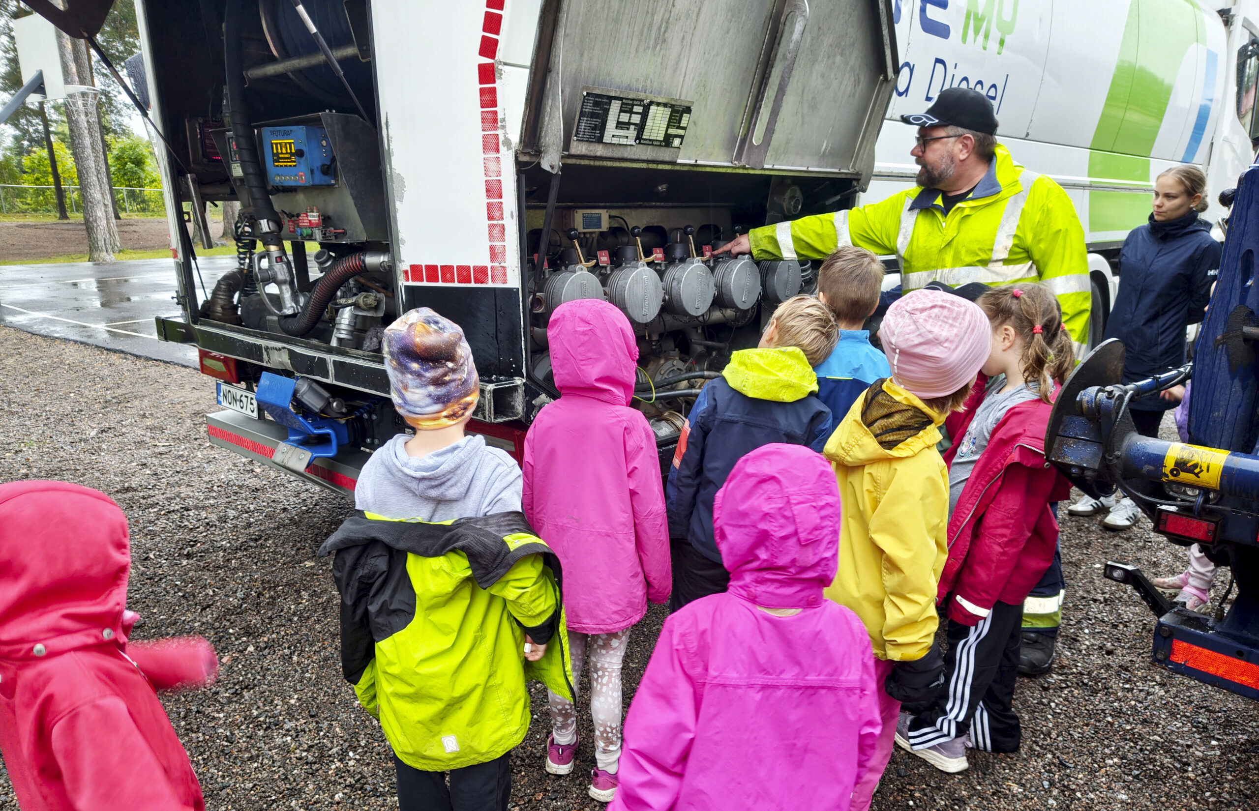 Lauma värikkäisiin vaatteisiin pukeutuneita lapsia seisoo säiliöauton edessä, jota esittelee henkilö neonvihreässä työtakissaan ja lippiksessään.