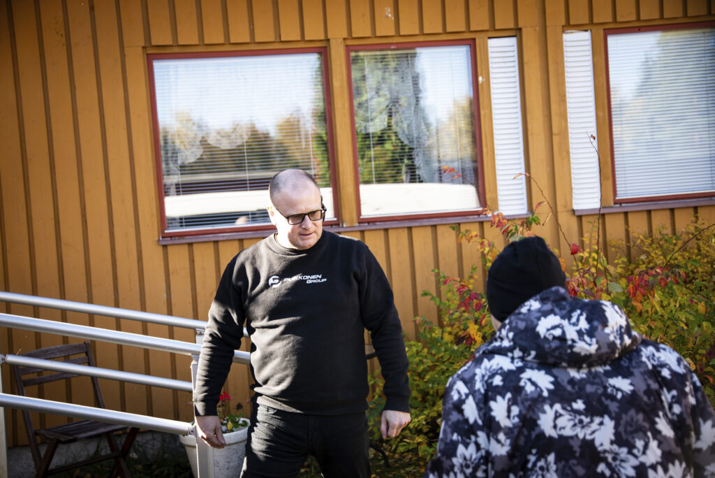 Mies ja ikäihminen mustavalkoharmaassa takissa keltaisen rakennuksen edustalla.