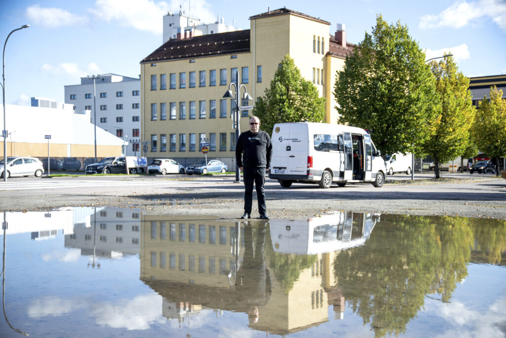 Mies seisoo heijastavan suuren lätäkön takana, joka heijastaa taustalla olevan näkymän sekä valkoisen pikkubussin ylösalaiseksi.