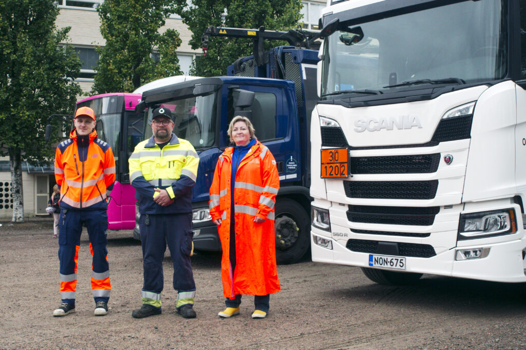 Kolme henkilöä kirkkaissa huomiotyövaatteissaan seisoo säiliöautojen edessä. 
