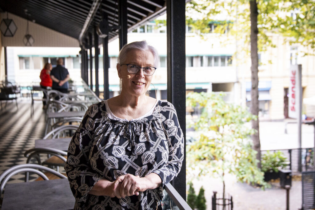 Silmälasipäinen nainen nojaa ravintolan parvekkeen kaiteeseen ja katsoo kameraan hymyillen.