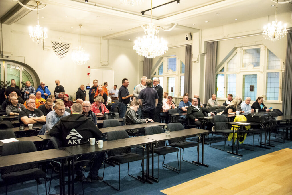 Tauolla olevia kurssille osallistujia tilassa, jossa pitkät mustat pöydät, sininen lattia, isot vanhat ikkunan ja isot kattokruunut.
