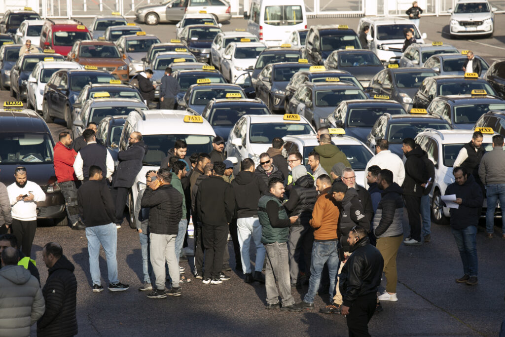Useita ihmisiä seisoo keskustellen parkkipaikalla jossa on pysäköitynä monta taksia, ja taustalla näkyy lisää ajoneuvoja.