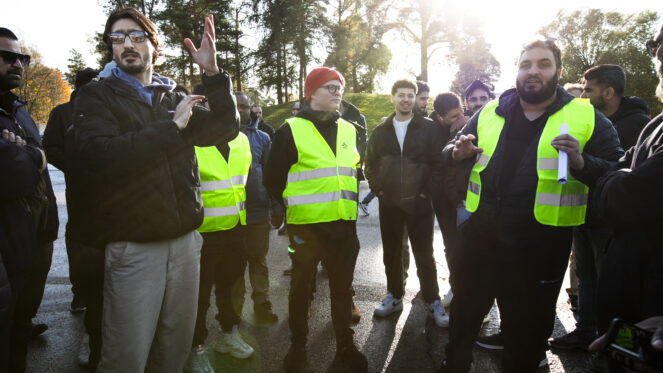 Ihmisiä seisoo ulkona auringonpaisteessa keltaisissa heijastinliiveissä ja keskustellen ja elehtien.