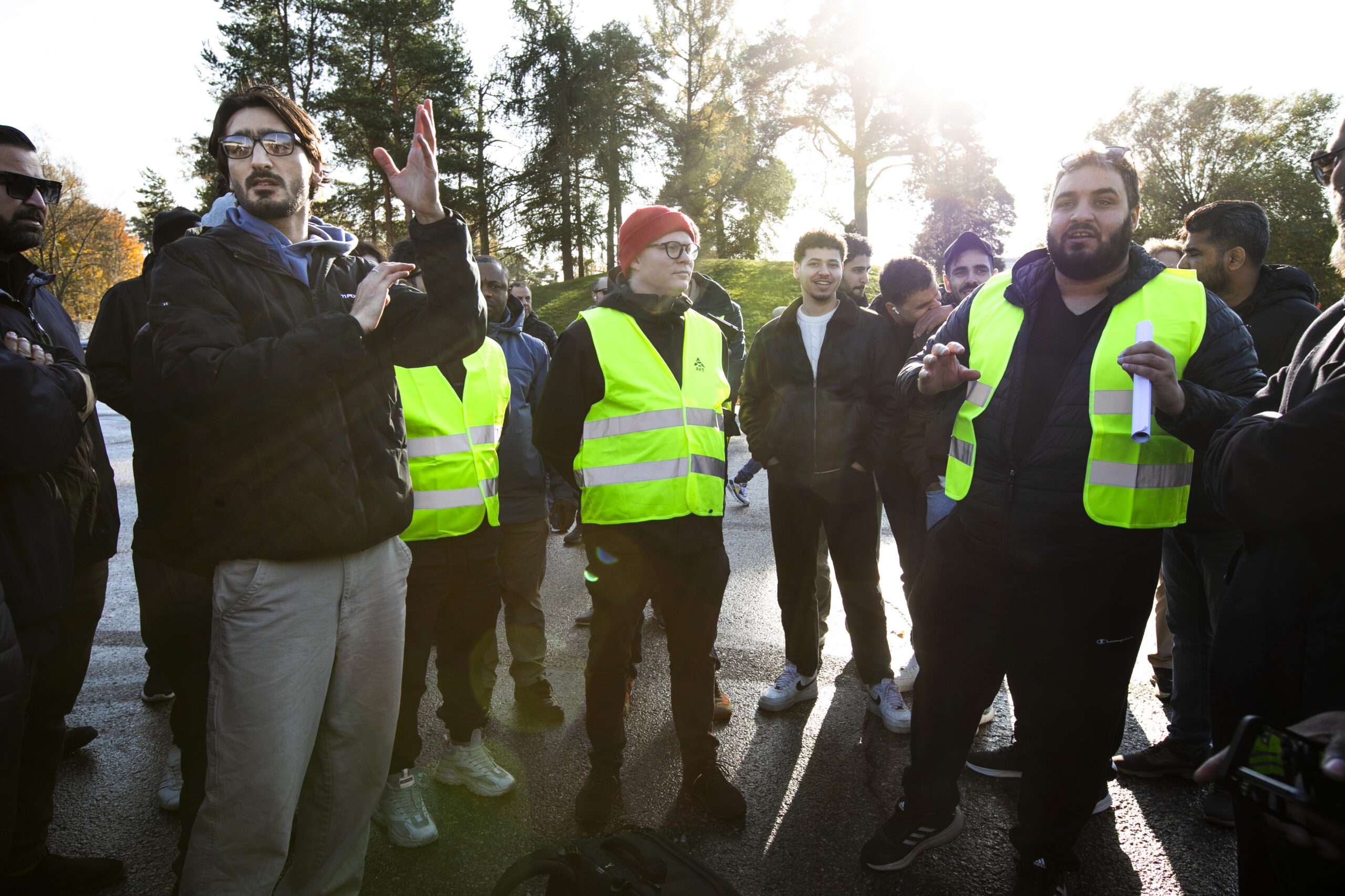 Ihmisiä seisoo ulkona auringonpaisteessa keltaisissa heijastinliiveissä ja keskustellen ja elehtien.