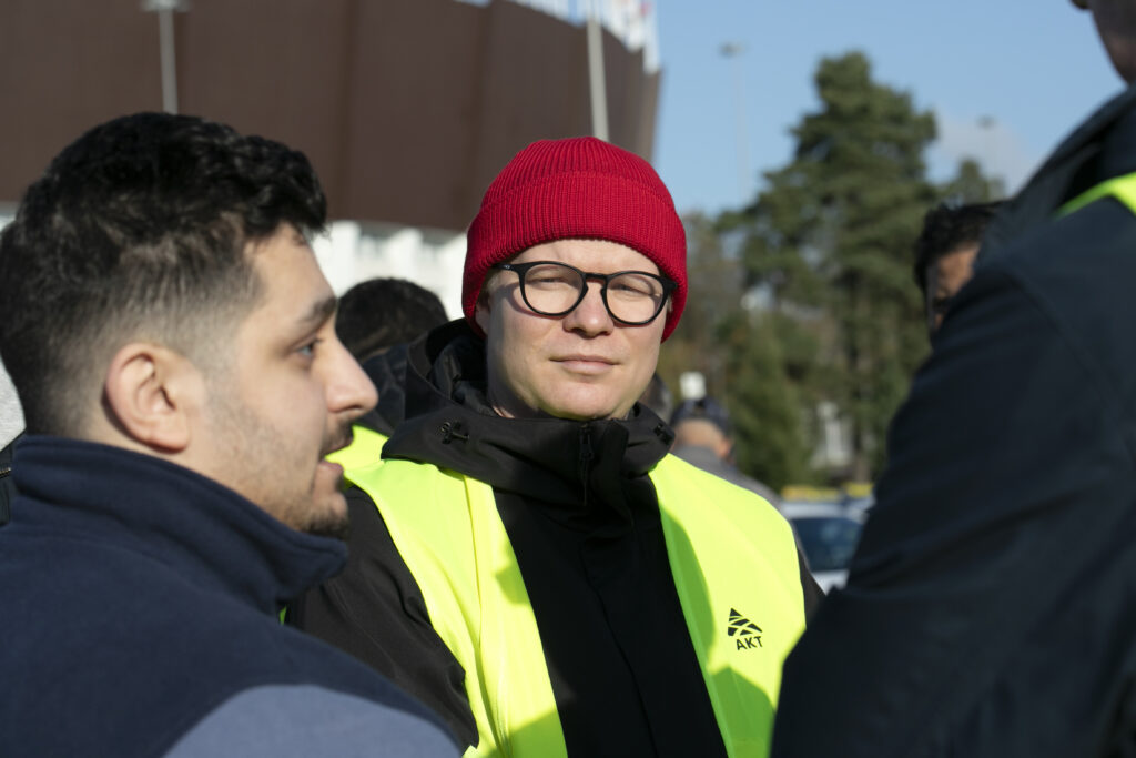 Kolme henkilöä keskustelee ulkona, joista yksi eli Niko Blom käyttää punaista pipoa, silmälaseja ja keltaista heijastinliiviä.

