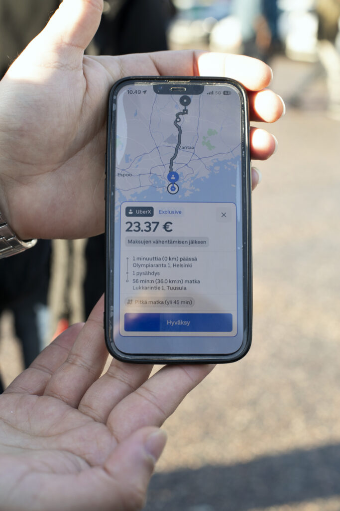 Henkilö pitelee kädessään puhelinta, jossa näkyy Uber-sovelluksen reittiohje 56 minuutin matkalle, jonka hinta on 23.37 €.