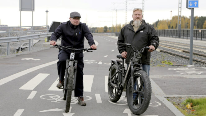 Två män står med sina cyklar på en cykelbana. En spårvagn närmar sig bakifrån.