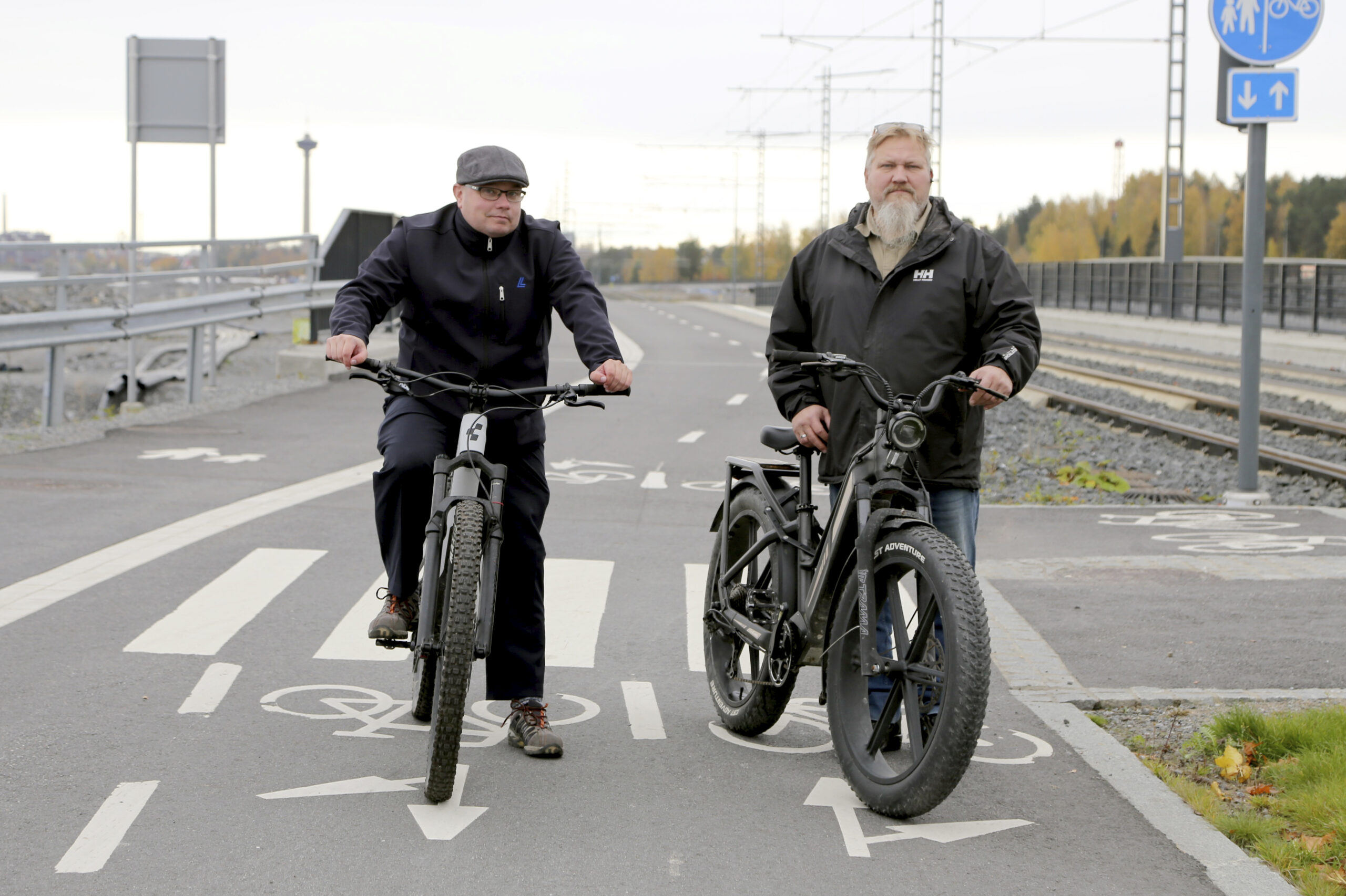 Två män står med sina cyklar på en cykelbana. En spårvagn närmar sig bakifrån.